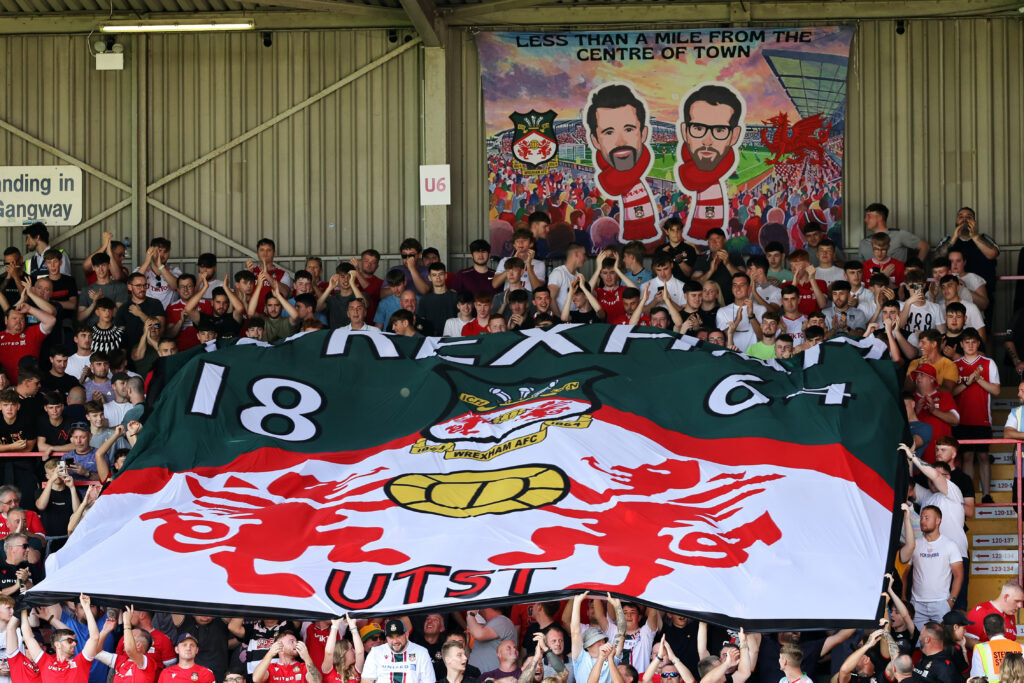 Fans of Wrexham display a flag of their badge with a banner of owners Rob McElhenney and Ryan Reynolds during the Sky Bet League Two match between Wrexham and Doncaster Rovers at Racecourse Ground on September 9, 2023 in Wrexham, Wales.